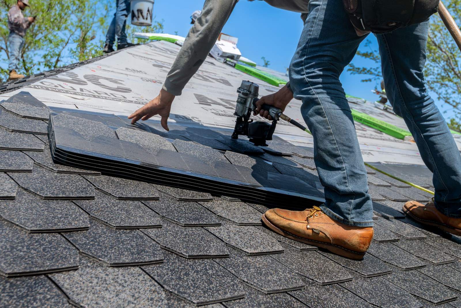 Roofer fastening asphalt shingles on a steep residential roof under a clear sky