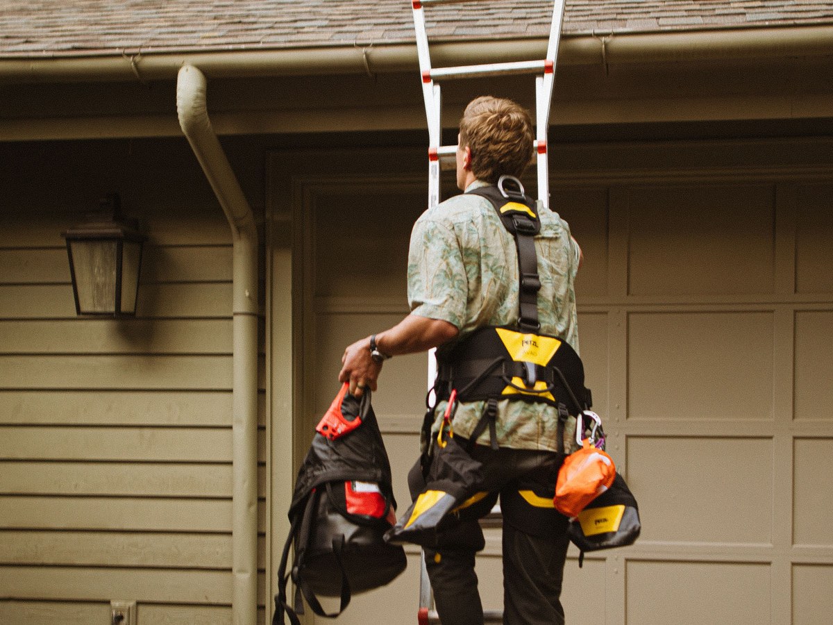 A roofer in a safety harness climbing a ladder to inspect a roof